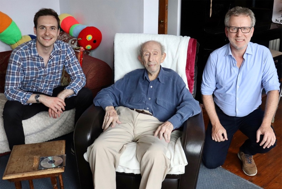 Three men; Eric Sanders on a reclining chair, he is wearing a blue shirt and a khaki trouser; two men on his either side; one is sitting on the sofa and the other sitting squatted on the floor.